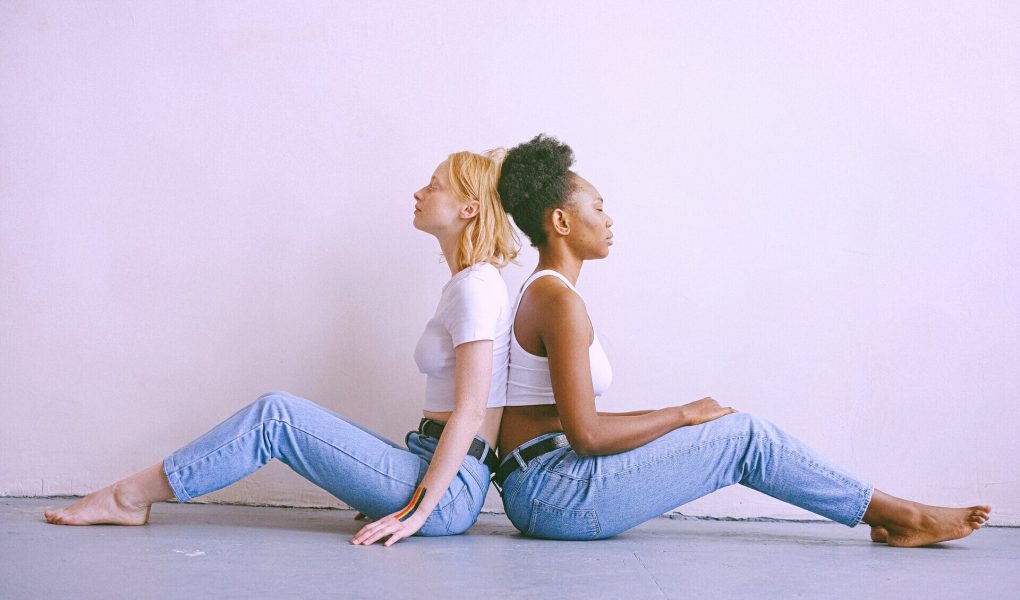 Zwei Frauen sitzen Rücken an Rücken vor einer hellen Wand. Sie tragen jeweils Jeans und ein weißes Top