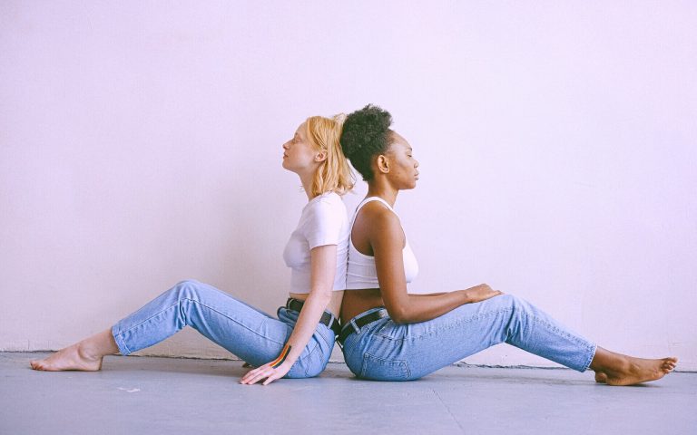 Zwei Frauen sitzen Rücken an Rücken vor einer hellen Wand. Sie tragen jeweils Jeans und ein weißes Top