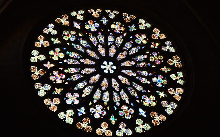 Ein rundes Buntglasfenster. Es sieht aus wie ein Mandala aus verschiedenen, kleinteiligen Formen bestehend aus buntem Glas