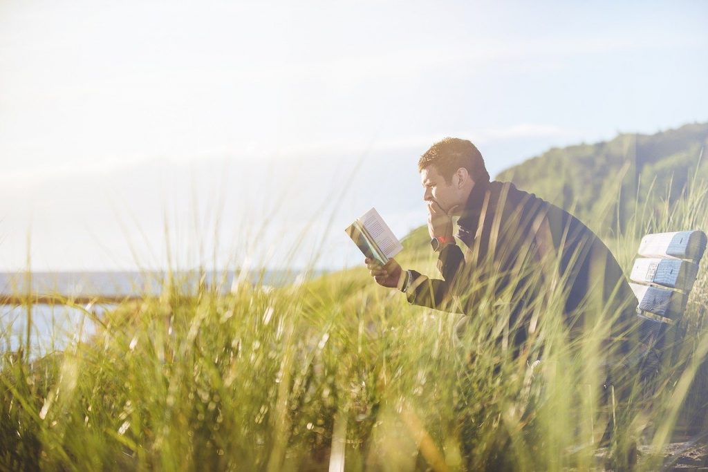 Ein Mann sitzt auf einer Bank und liest konzentriert in einem Buch. Um ihn herum ist viel hohes Gras, im Hintergrund sieht man das Meer.