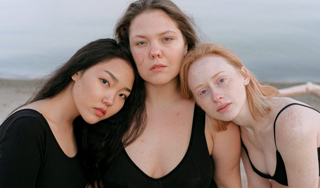 Drei Frauen schauen in die Kamera. Ihre Köpfe sind aneinander gelehnt. Im Hintergrund sieht man einen Strand. Eine hat schwarze haare, eine braune und eine rötliche.