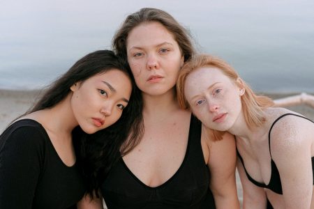 Drei Frauen schauen in die Kamera. Ihre Köpfe sind aneinander gelehnt. Im Hintergrund sieht man einen Strand. Eine hat schwarze haare, eine braune und eine rötliche.