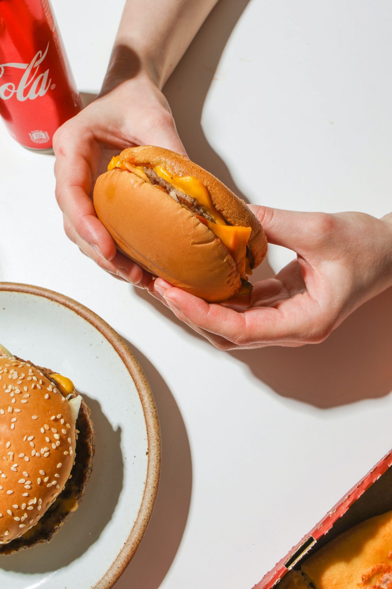Ein Paar Hände hält einen Burger, aus dem der geschmolzene Käse läuft. In der linken unteren Ecke sieht man etwa die Hälfte eines Tellers mit einem weiteren Hamburger darauf. In der linken oberen Ecke steht eine Getränkedose.
