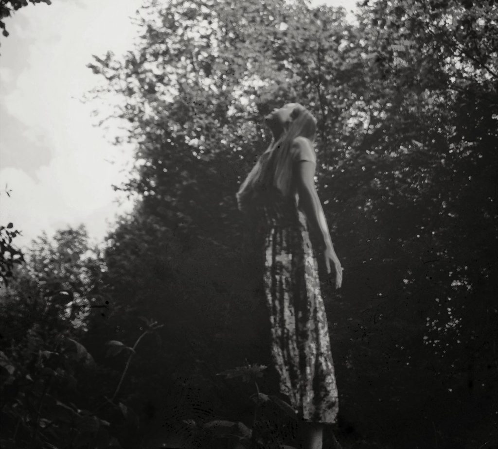 Das Bild ist schwarz-weiß. Ein Mädchen in einem gemusterten Rock steht im Wald und sieht nach oben. Ihre Hand liegt auf ihrer Brust