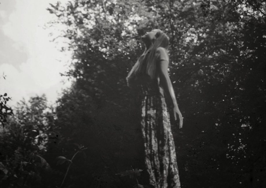 Das Bild ist schwarz-weiß. Ein Mädchen in einem gemusterten Rock steht im Wald und sieht nach oben. Ihre Hand liegt auf ihrer Brust
