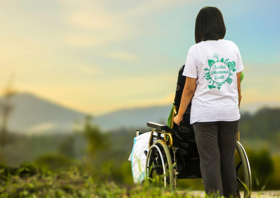 Eine junge Frau schiebt einen Rollstuhl. Zusammen schauen sie über ein grünes Tal