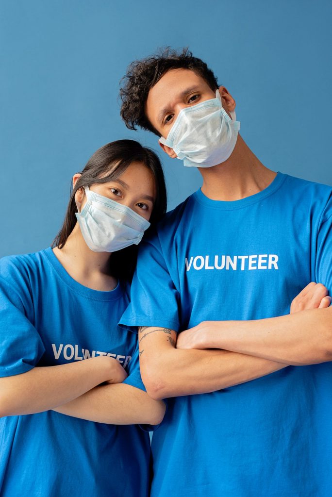 Zwei dunkelhaarige junge Menschen stehen nebeneinander mit verschränkten Armen, lehnen sich aneinander und schauen in die Kamera. Sie tragen blaue T-Shirts mit der Aufschrift "Volunteer"